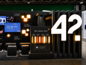 Exhibition booth for the brand 42, featuring black walls, digital screens displaying images, orange-illuminated panels, and a large "42?" sign prominently displayed on the right.