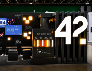 Stand de exposição da marca 42, com paredes pretas, telas digitais exibindo imagens, painéis iluminados em laranja e um grande número "42?" em destaque à direita.